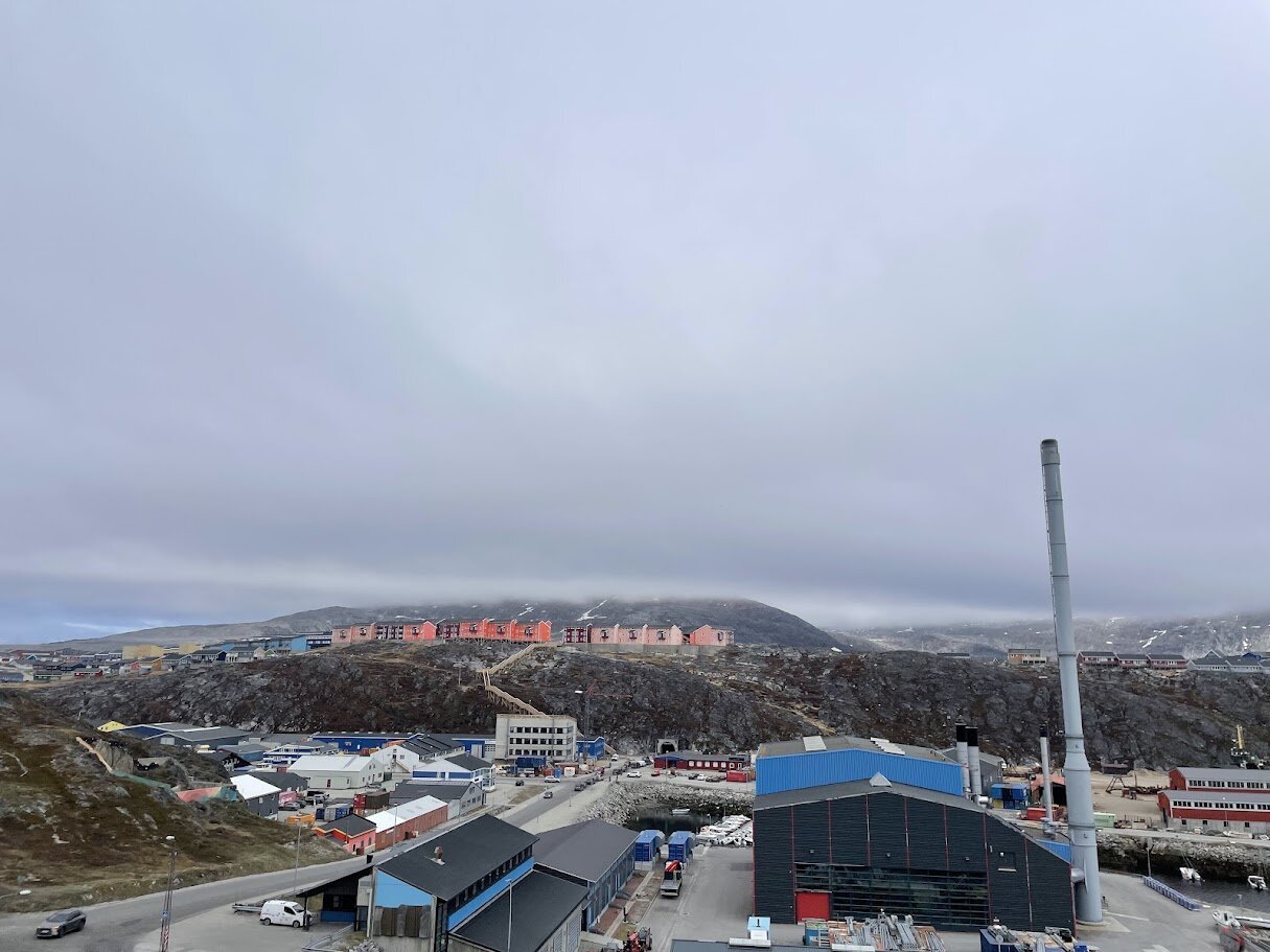 Udsigt over Nuuk med farverige boligblokke på fjeldsiden og industrihavnen under en grå himmel.