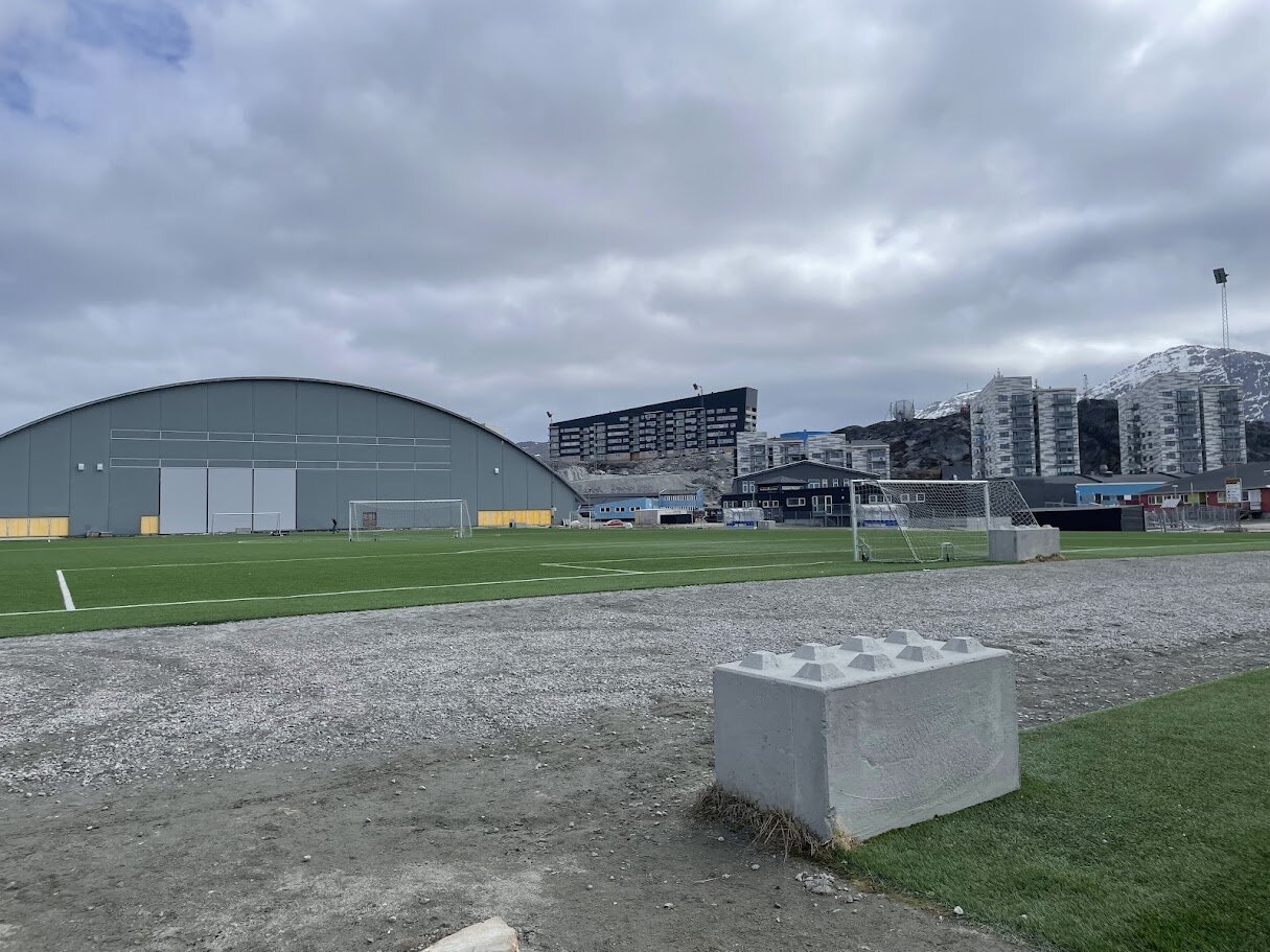 Nuuks fodboldstadion med kunstgræs, Godthåbhallens buede tag og boligblokke under fjeldet.