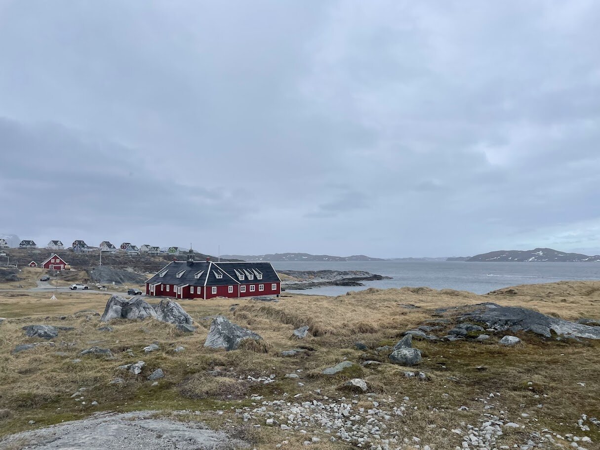 Rødt træhus ved den græs- og klippeklædte kyst med fjorden og Nuuks huse i baggrunden.
