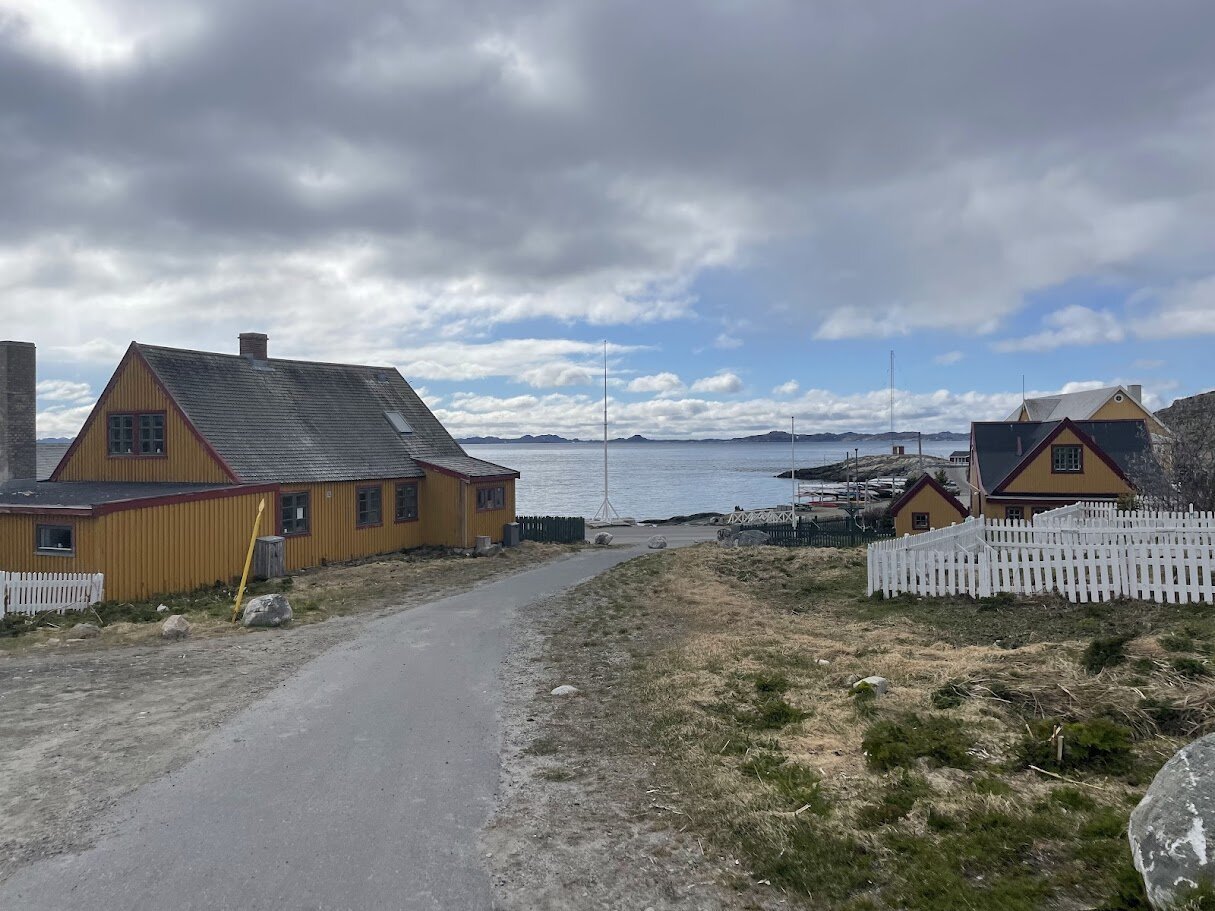 Gult træhus med rødt tag i den gamle kolonihavn, en grusvej fører ned mod fjorden.