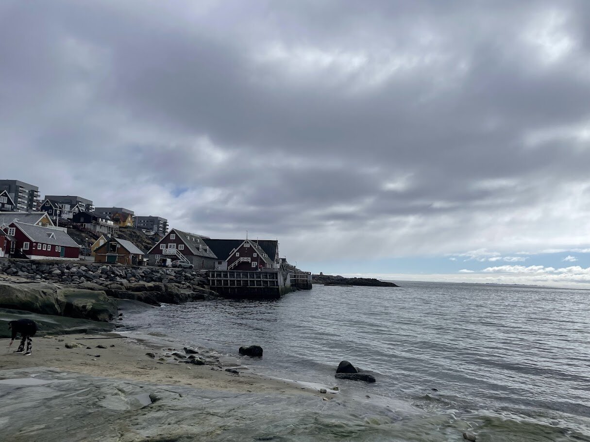 Række af farverige træhuse langs vandet i den gamle kolonihavn med klipper i forgrunden.